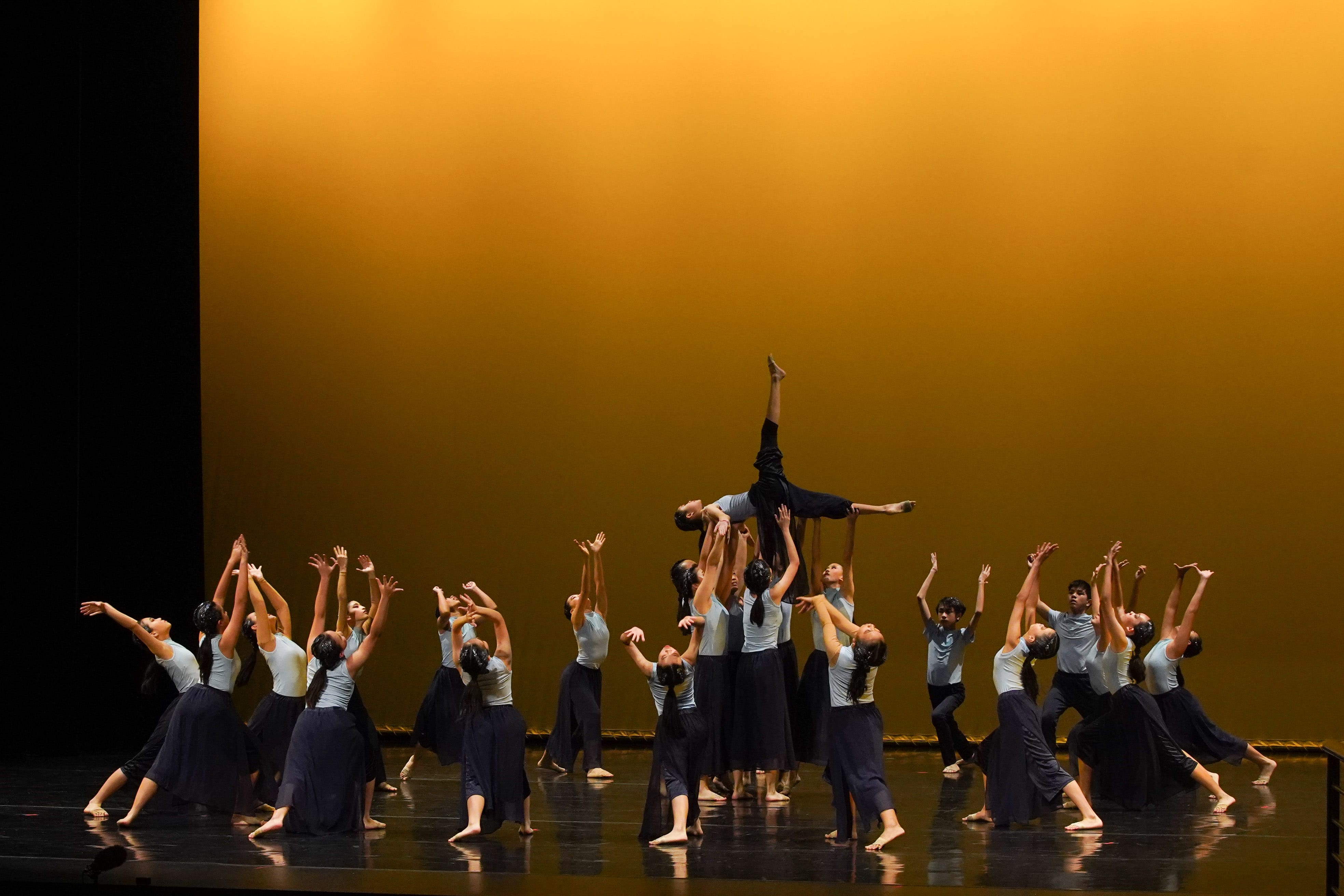 Large group of dancers on a lit stage lifting one performer overhead against a golden backdrop.